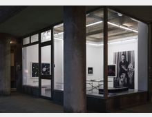 Straßenansicht einer Galerie mit großen Glasfenstern, durch die eine minimalistische Ausstellung sichtbar ist. Im Innenraum sind mehrere Bilderrahmen mit Kunstwerken an weißen Wänden sowie ein Tisch mit Prospekten und Bänken zu sehen; ein großes Schwarz-Weiß-Porträt dominiert die rechte Wand.