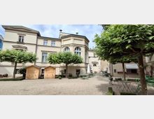 Innenhof vor einem hellen, klassizistischen Gebäude mit großen Sprossenfenstern und einem vorspringenden Erker mit Rundbogenfenstern. Kiesfläche mit beschnittenen Bäumen, verstreuten Bistro-Tischen und -Stühlen, Sonnenschirmen sowie zwei kleinen Holzbuden.