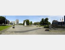 Weitläufiger Platz mit gepflastertem Boden, einer zentralen Wasserfontäne und Bänken im Vordergrund. Umgeben von modernen Bürogebäuden, vereinzelten Bäumen und wehenden Flaggen in einer urbanen Umgebung.