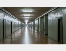 Langer, geradliniger Flur mit glänzenden, hellgrünen Wänden, hölzernem Boden und Deckenleuchten in Reihen. Beidseitig befinden sich mehrere geschlossene Türen, teilweise mit Glasfenstern, sowie Feuerlöscher an den Wänden.