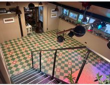 Innenraum mit einem bunt gemusterten Boden aus grün-weißen Fliesen und roten Akzenten, gesehen von einer Metalltreppe mit Geländer aus Drahtseilen. An der Decke hängen mehrere schwarze Lampen, und auf einer Fensterbank stehen zahlreiche Pflanzen.