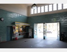 Eingangsbereich eines Gebäudes mit türkisfarbenen Fliesenwänden, einem kleinen Kiosk für Zeitungen, Getränke und Süßwaren sowie Glastüren, die nach außen führen. Oberhalb der Türen sind rechteckige Fenster, durch die Tageslicht einfällt.