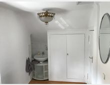 Ein kleiner Raum mit weiß gestrichenen Wänden, einer runden Spiegelfläche an der rechten Wand, einem Waschbecken auf einem Eckschrank mit Ablageflächen und Handtüchern an der linken Seite. An der Rückwand befindet sich ein weißer Einbauschrank, und die Decke ist mit einem Kronleuchter mit Kristallelementen geschmückt.