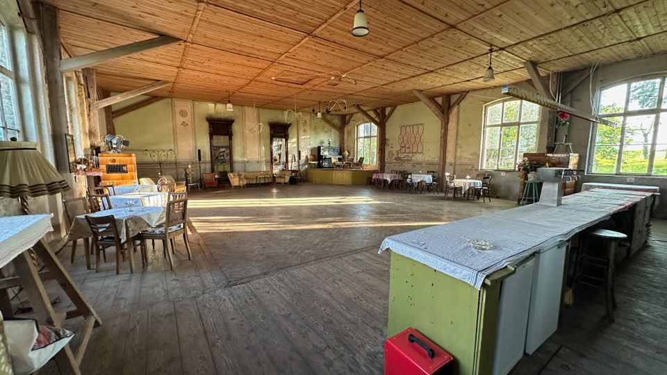 Großer, rustikaler Saal mit Holzfußboden und Holzdecke, sichtbaren Stützbalken und mehreren hängenden Lampen. Entlang der Wände stehen kleine Tische und Stühle, im Vordergrund eine lange Theke; große Sprossenfenster lassen viel Tageslicht herein und geben den Blick ins Grüne frei.