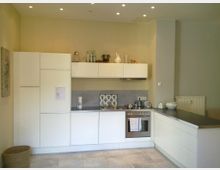 Moderne, helle Küche mit weißen Einbauschränken, einem Herd mit Edelstahl-Backofen, einer grauen Arbeitsplatte und neutralen Fliesenboden. An der Wand befinden sich Deko-Gegenstände und ein Heizkörper ist sichtbar.