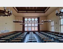 Großer Hörsaal mit hohen Decken, mehreren runden Kronleuchtern und langen Reihen aus Tischen und Stühlen. An der Stirnseite befindet sich ein großes Fenster mit runden Oberlichtern, seitlich hohe Fenster und an den Wänden Informationstafeln.