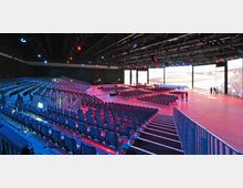 Großer Veranstaltungssaal mit einer ansteigenden Tribüne aus Sitzreihen auf der linken Seite, freien Stuhlreihen auf einer flachen Ebene rechts und mehreren großen Bildschirmen an der Rückwand. Die Decke ist mit Scheinwerfern und Bühnentechnik ausgestattet, der Saal ist in farbiges Licht getaucht.