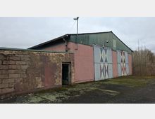 Außenansicht einer einfachen Lager- oder Industriehalle mit grünem Wellblechdach und rosafarbenen Wänden, davor ein kleiner Anbau mit verwitterter Fassade und offener Tür. Große Metall-Schiebetore mit geometrischen X-Mustern und ein Lampenmast stehen vor einem asphaltierten Hof, im Hintergrund sind kahle Bäume und ein grauer Himmel zu sehen.