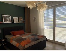 Modernes Schlafzimmer mit großem Polsterbett und karierter Bettdecke vor einer dunkelgrünen Akzentwand mit Bilderleiste. Rechts bodentiefe Fenster mit Lamellenjalousien und transparenten Vorhängen; an der Decke eine wolkenartige Hängeleuchte.