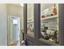 Kleine Küche mit weißen Wandfliesen, offener Regalablage und verschiedenen Küchenutensilien. Im Vordergrund sind eine Espressomaschine und andere Gerätschaften zu sehen, während eine Tür mit Milchglasfenster in einen angrenzenden Raum führt.