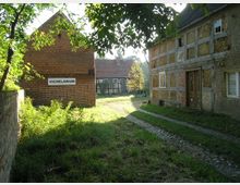 Ein rustikaler Hof mit mehreren historischen Gebäuden, darunter ein Ziegelgebäude mit der Aufschrift „Vichelarium“ und ein Fachwerkhaus mit sichtbaren Lehmwänden. Der Vordergrund zeigt einen grasbewachsenen Weg, der von Bäumen und Sträuchern gesäumt ist.