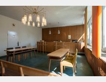 Ein Büroraum mit Holzmöbeln im Retro-Stil, bestehend aus einem großen Holztisch mit Stühlen, einer hölzernen Empfangstheke und Wandverkleidungen. Große Fenster auf der rechten Seite lassen Tageslicht herein, während ein Kronleuchter mit mehreren Leuchten die Decke schmückt.