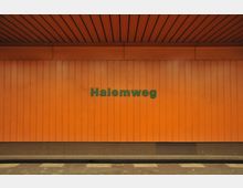 Orange Wandverkleidung in einer U-Bahn-Station mit dem Schriftzug „Halemweg“ in grünen Buchstaben. Der Bodenbereich zeigt das Gleis und den Bahnsteig.