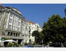 Historisches Gebäude mit dekorativer Fassade und Säulen im unteren Bereich, daneben ein helles Wohnhaus mit Balkonen, beide mit roten Ziegeldächern. Im Vordergrund sind parkende Autos, Bäume und ein Straßencafé zu sehen, der Himmel ist klar und blau.