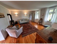 Helles Wohnzimmer mit Holzboden, einer grauen Couch, einem beigen Sessel und einem Kaminofen auf der linken Seite. Ein Teppich mit orientalischem Muster liegt in der Mitte, während zwei Fenster mit weißen Vorhängen Tageslicht einlassen.