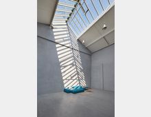 Moderner Raum mit hohen Wänden aus grauem Beton, beleuchtet durch ein großes Schrägdachfenster, das Schattenlinien wirft. Im Vordergrund befindet sich ein blaues Sofa auf einem kleinen Teppich, ergänzt durch einen runden Holztisch.