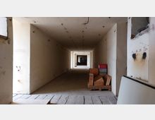 Langer, unbelebter Gang mit beigen Wänden und sichtbaren Schäden, der zu einem weiteren Raum mit Glasfenstern führt. Rechts sind nummerierte Wandmarkierungen und gestapelte Holzplatten sowie rote Kunststoffboxen auf einer Palette sichtbar.