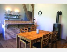 Wohnzimmer mit Essbereich in rustikalem Stil: Ein Holztisch mit passenden Stühlen steht auf einem Holzboden, daneben befindet sich ein moderner Kaminofen. Im Hintergrund sind eine Theke aus Ziegelsteinen, offene Regale mit Geschirr und eine Wanduhr zu sehen.