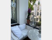 Eine kleine Balkon-Sitzecke mit einer weißen gepolsterten Bank, einem runden Steintisch und einer Topfpflanze mit lila Blüten. Im Hintergrund sind ein Geländer, eine Glaslaterne und die Fassade eines weißen Altbaugebäudes mit verzierten Fenstern zu sehen.