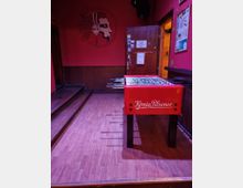 Ein roter Tischkicker mit der Aufschrift "König Pilsener" steht auf einem Holzboden in einem Innenraum mit roter Wandfarbe. Im Hintergrund ist eine Holztür mit Aushängen sowie ein Wandbild eines Gesichts mit Kopfhörern zu sehen.