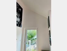 Innenraum mit hoher, weißer Wand und einem großen Fenster, das auf eine Terrasse mit Sitzmöbeln und einen Garten mit Bäumen und Pflanzen blickt. An den Wänden hängen moderne Kunstwerke, und draußen ist ein Teil eines Dachvorsprungs sichtbar.