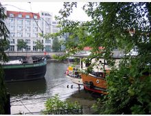 Zwei Boote liegen an einem bewucherten Flussufer, umgeben von Bäumen. Im Hintergrund sind mehrstöckige, moderne Gebäude mit weißen Fassaden und roten Dächern zu sehen.