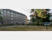 Wohnsiedlung mit vierstöckigen Mehrfamilienhäusern aus Beton, umgeben von Grünflächen und Bäumen. In der Mitte befindet sich ein Freiraum mit Wäscheleinen und einem Gehweg.