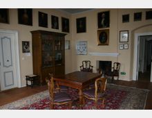 Historischer Raum mit Holzmöbeln, darunter ein rechteckiger Tisch und Stühle mit gestreiften Polstern, auf einem gemusterten Teppich. An den Wänden hängen zahlreiche gerahmte Porträts und ein Bücherschrank steht neben einem Kamin.