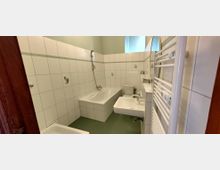 Kleines, gefliestes Badezimmer mit Badewanne und Duscharmatur an der Rückwand, WC und wandhängendem Waschbecken. Rechts befindet sich ein großer Handtuchheizkörper, darüber ein Fenster für Tageslicht.