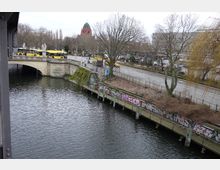 Blick auf eine steinerne Brücke über einen Fluss, mit einer Uferpromenade, die mit Graffiti bedeckt ist. Im Hintergrund sind eine Straße mit Bäumen, gelbe Busse und ein rotes Kirchengebäude mit Kuppel zu sehen.