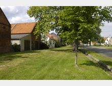 Dorfstraße mit einer Reihe von Häusern, teils aus Backstein und teils verputzt, und roten Ziegeldächern auf der linken Seite sowie Bäumen und einer Rasenfläche entlang der Straße. Rechts verläuft eine asphaltierte Fahrbahn mit Gehwegen.