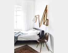 Bright minimalist bedroom with a low gray upholstered bed dressed in white bedding beside a large window with sheer white curtains. A wooden accordion wall-mounted lamp extends over the bed, and a molded chair and patterned rug sit on white-painted floorboards.
