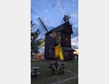 Hölzerne Windmühle mit hohem, dunkel verkleidetem Turm und großen Flügeln, von warmen Lichterketten umspannt, vor einem Abendhimmel mit Wolken. Im Vordergrund stehen Rasenfläche und rustikale Holzelemente, im Hintergrund weitere Gebäude auf dem Gelände.