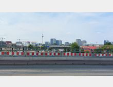 Stadtansicht mit einem erhöhten Blick auf eine Baustelle im Vordergrund, hinter der sich moderne Hochhäuser und der Fernsehturm vor einem blauen Himmel erheben. Im Mittelgrund sind kleinere Gewerbegebäude und Werbeschilder sichtbar.