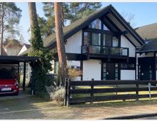 Zweistöckiges Einfamilienhaus mit schwarzem Satteldach und weißer Fassade, ergänzt durch große Glasfenster und einen Balkon. Im Vordergrund befindet sich ein Carport mit einem roten Auto, während der Bereich von Bäumen und einem Holzzaun umgeben ist.