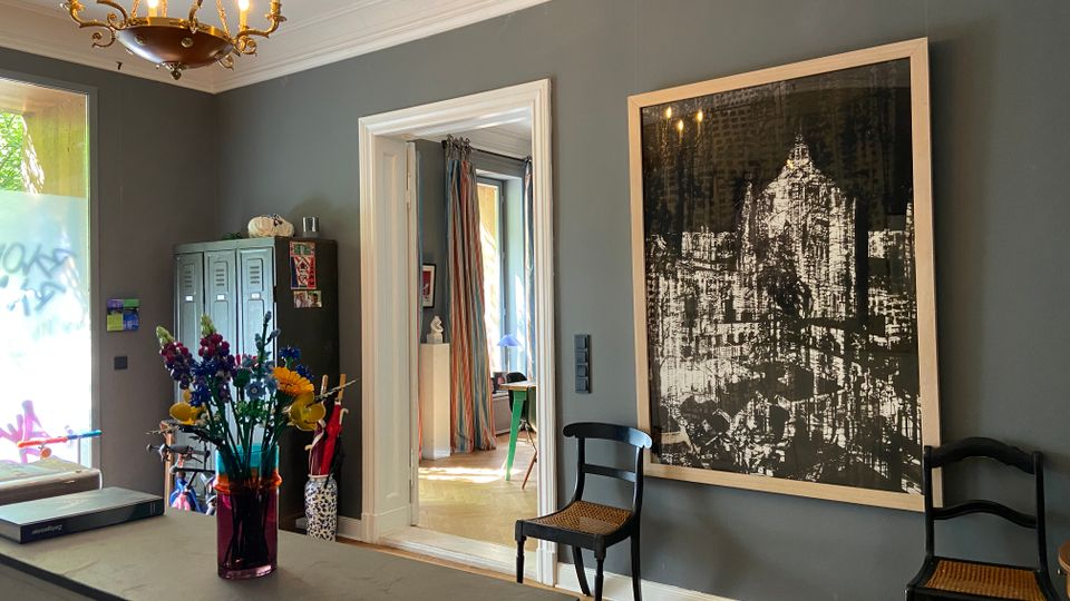 Innenansicht eines Wohnraums mit grauen Wänden, Stuckdecke und einem Messing-Kronleuchter; an der Wand hängt ein großes gerahmtes Schwarzweißbild einer Stadtansicht neben zwei Stühlen. Links stehen ein Metallschrank und ein Tisch mit Blumen, und durch eine Türöffnung ist ein weiterer Raum mit Vorhängen und Fenster zu sehen.