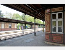 Bahnhof mit überdachten Bahnsteigen aus Backstein und dekorativen Mustern an den Wänden. Im Hintergrund sind Gleise, Schilder und grüne Bäume zu sehen, während ein Fenster und eine Säule im Vordergrund stehen.