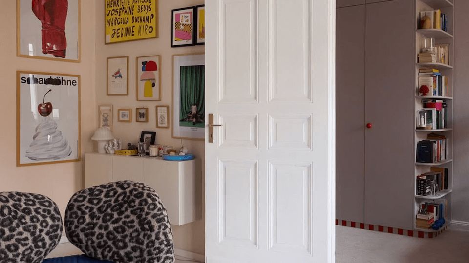 Eclectic apartment living room with white-painted floors, ornate ceiling molding, and a tall white paneled door opening into an adjoining room. The walls are covered with framed art and a red lips wall sculpture, with blue seating, patterned cushions, and shelving visible through the doorway.