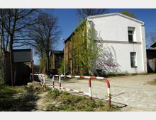 Eine gepflasterte Seitenstraße mit einer rot-weißen Absperrung im Vordergrund, flankiert von einem weiß verputzten Gebäude mit Graffiti und einem backsteinernen Haus. Im Hintergrund sind Bäume und parkende Autos zu sehen, unter einem klaren blauen Himmel.