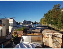 Ein kleiner Fluss mit geparkten Booten, darunter ein Hausboot mit runden Fenstern und einer offenen Tür. Auf dem Deck befinden sich Pflanzkästen, und am Ufer wachsen dichte Bäume unter klarem, blauem Himmel.