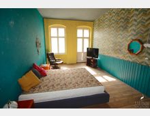 Ein schlicht eingerichtetes Zimmer mit einer bunten Wandgestaltung: eine blaue Wand, eine gelbe Wand und eine gemusterte Tapete in Blau-Gelb-Tönen. Im Raum stehen ein Bett mit geblümtem Bezug, ein Sessel, ein TV-Gerät auf einem kleinen Tisch sowie ein Holzfußboden und große Fenster, die viel Licht hereinlassen.