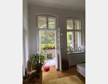 Wohnzimmer mit Holzboden und geöffneten Doppelfenstern, die zu einem kleinen Balkon führen, der mit Pflanzen, einem Sitzkissen und Dekorationen geschmückt ist. Im Innenraum stehen eine Topfpflanze auf einem Regal und ein weißer Heizkörper unter weiteren Fenstern.