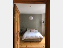 Schlafzimmer mit einem Doppelbett in der Mitte, weißer Bettwäsche und einem pastellrosa Kissen. Der Raum hat Holzböden, eine graue Wand, eine hängende Zimmerpflanze und eine weiße Pendelleuchte.