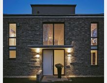 Moderne Backsteinfassade eines Wohnhauses bei Dämmerung, mit einer zentralen weißen Haustür, flankiert von beleuchteten Wandleuchten. Große Fenster auf beiden Seiten und darüber lassen Innenlicht nach außen strahlen, ein schwarzer Pflanzkübel steht vor der Tür.