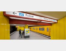 U-Bahnhof mit gelben Wänden und einem durchfahrenden gelben Zug der Linie U7. Ein Wegweiser zeigt die Richtungen "U7 Rudow" und "Rathaus Spandau", während Fahrgäste auf der Plattform warten.