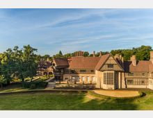 Luftansicht eines historischen Gebäudes im Fachwerkstil mit roten Ziegeldächern, großen Fenstern und Verzierungen. Das Gebäude liegt inmitten einer grünen Landschaft mit angrenzendem Wald und einem gepflegten Garten.
