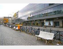 Straßenszene mit einem modernen Gebäude mit großen Glasfassaden im Hintergrund, einem Gehweg mit Kopfsteinpflaster, aufgereihten Sitzbänken, angeschlossenen Fahrrädern und einer Haltestelle mit einem gelben Linienbus. Einige Fußgänger, Tauben und Laternen vervollständigen das Stadtbild.