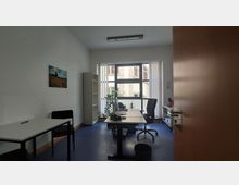 Kleines Büro mit Schreibtisch, Bürostuhl und Computer am Fenster. Links steht ein zusätzlicher Tisch mit einem Stuhl, ein Regal sowie Pflanzen und Lamellenjalousien ergänzen den Raum.