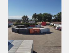 Eine Industriefläche mit aufgestapelten Schiffscontainern in unterschiedlichen Farben, umgeben von Bäumen und Gebäuden. Im Hintergrund sind Felder, ein Wohnhaus und weitere Container zu sehen.