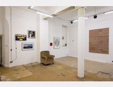 Ein minimalistischer Ausstellungsraum mit weißen Wänden, einer einzelnen Säule und freiliegendem, abgenutztem Boden. An den Wänden hängen mehrere Kunstwerke, darunter Fotografien, eine Neon-Tafel mit der Aufschrift „I want to be an artist too“ und ein großformatiges Streifenbild; links steht ein brauner Polstersessel.